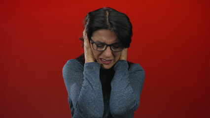 Middle age hispanic woman grimacing with pained expression while covering ears in red studio;...