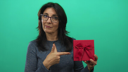 Middle age hispanic woman pointing finger to red gift box held at chest level in a green studio backdrop; celebration.