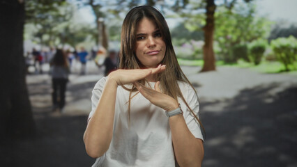 Young hispanic woman in park gesturing timeout while surrounded by vibrant greenery and blurred people in background conveying outdoor leisure and relaxation.