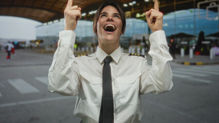 Woman pilot smiling in uniform pointing upwards at airport outdoor showcasing enthusiasm and pride in aviation profession outside terminal building background.