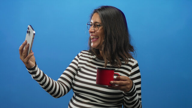 Woman holds smartphone in one hand and red mug in the other, hand raised to take a selfie in a studio with blue backdrop; amused connection.