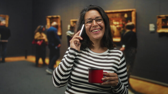 Woman holds phone to ear and a red mug while puckering lips in a museum gallery beside framed paintings and visitors; playful moment.