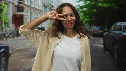 Woman making a peace sign with fingers at her eye while smiling in an urban street scene with parked cars and bicycles visible; playful joy.