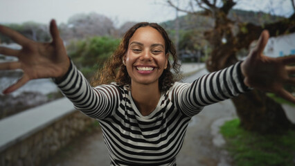 Woman with arms extended reaching toward camera on street promenade by the seaside, smiling with eyes closed; joyful embrace.