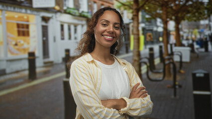 Young african american woman smiling with arms crossed, wearing a striped shirt on a city street; confidence.