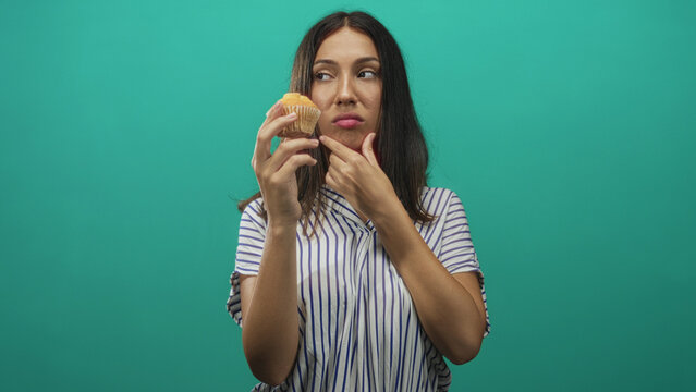 Young hispanic brunette woman holding a muffin while touching chin with hand in teal studio, striped shirt visible; doubt.