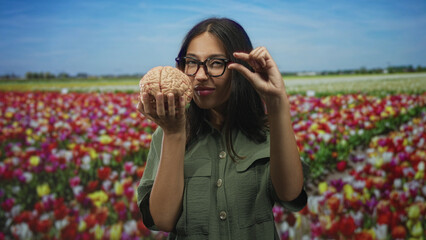 Young brunette woman holding model brain with pinching fingers in colorful tulip field, smiling  curiosity. © Krakenimages.com