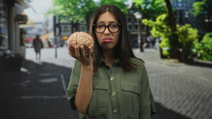 Fototapeta premium Woman holding a brain model at eye level on a street in the city, wearing glasses and an olive shirt; confusion.