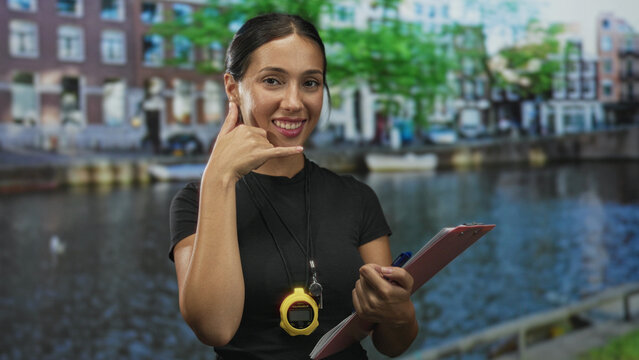 Young brunette woman trainer holds clipboard and yellow stopwatch and smiles, points finger at camera near canal and brick building on street; confidence leadership.