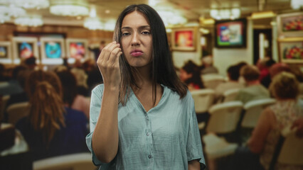 Woman young brunette pinching fingers near face with raised hand in gallery building among seated visitors; annoyance disapproval.