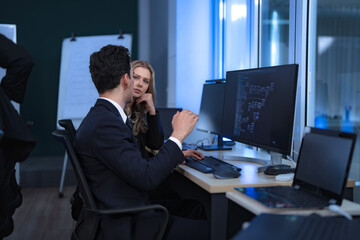 A diverse team of professionals collaborates in a modern tech office at night. A male and female colleague have a positive discussion, while others brainstorm at a whiteboard in the background.