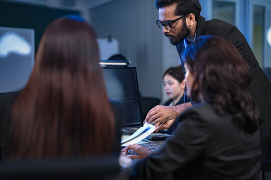 A senior data scientist with glasses points out an anomaly in a machine learning model's code to two female AI engineers, as they finalize the algorithm for a new predictive analytics service.