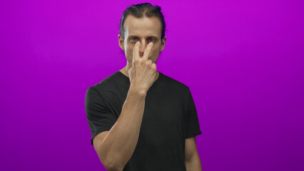 Young hispanic man with long hair holding two fingers to his face and pointing at camera in purple studio; confidence.