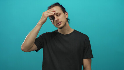 Young hispanic man with long hair, hand on forehead and eyes closed against a teal studio backdrop;...