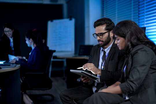 A diverse team of professionals collaborates on a business strategy late at night. A male manager uses a tablet and stylus to explain key points to his female colleague in a modern boardroom.