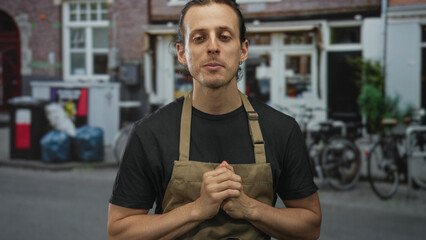 Young hispanic man with long hair and hands crossed over chest wearing a tan apron and black t shirt standing on a street outside a cafe; gratitude humility.