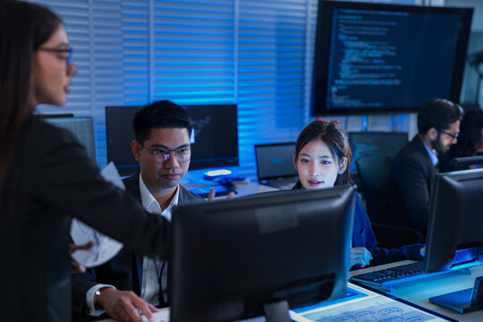 A female project manager leads a late-night review with her diverse team, discussing a report and analyzing data on computer screens to ensure they meet a critical deadline.