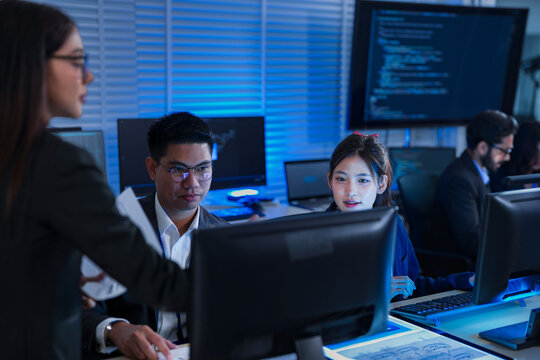 A female project manager leads a late-night review with her diverse team, discussing a report and analyzing data on computer screens to ensure they meet a critical deadline.