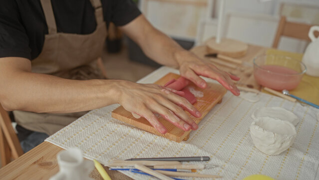 Man rolling pink clay with hands and wooden roller on a workboard in artist studio surrounded by brushes and bowls; concentration.