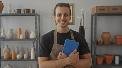 Man holding tablet to chest in pottery studio among shelves of handmade ceramics, vases and terracotta pots wearing an apron and smiling; craft pride.