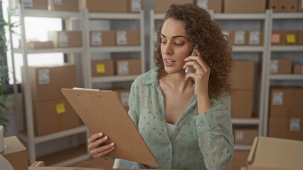 Woman holding clipboard and phone to ear while reading order details in building; focused...