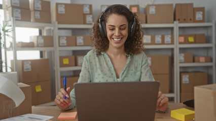 Woman wearing headphones smiling and gesturing with pen at laptop amid parcels on shelves in...