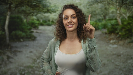 Woman points index finger up on forest path with bare chest visible in white tank top; assertion...