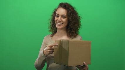 Woman smiling and holding a cardboard box while pointing to the package in a green studio; cheerful delivery.
