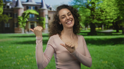 Woman smiling and points thumb to building beside castle in park with hands visible and relaxed posture; invitation confidence joy.