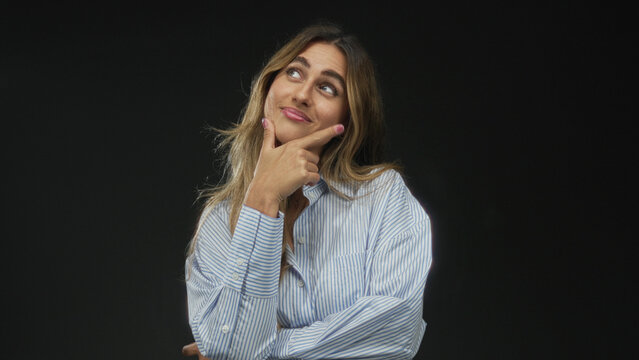 Woman holding chin with hand while gazing upward against a black studio background; thought curiosity introspection reflection.
