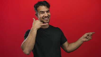 Young man gesturing call me with a playful expression against an isolated red background.