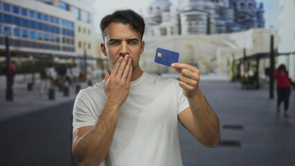 Young hispanic man on city street outdoors holds credit card with a surprised look on his face,...