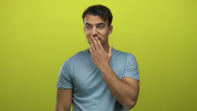 Young hispanic man wearing blue shirt over isolated yellow background appears surprised while covering mouth with hand, conveying shock and disbelief.