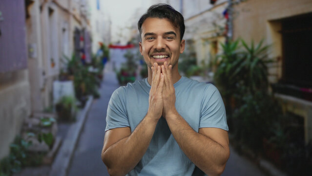 Young hispanic man in blue shirt smiling on a vibrant street, surrounded by plants and bright urban scenery during daylight, conveying a positive outdoor atmosphere.