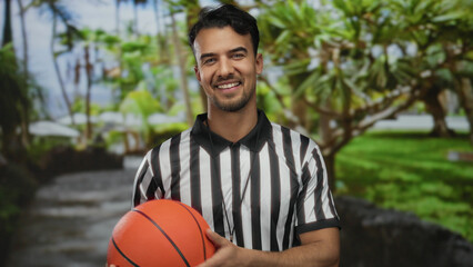 Hispanic man in referee uniform holding basketball in a vibrant park setting, smiling confidently with lush greenery in the background, showcasing an outdoor sports vibe.