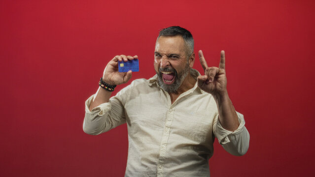 Man holding blue creditcard up to camera in red studio while smiling and making rock horns; financial confidence. - Powered by Adobe
