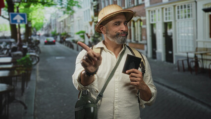 Man holding passport and pointing index finger while wearing explorer hat on a city street, backpack visible and canteen at hip; curiosity.