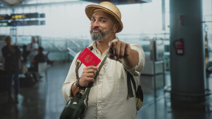 Middle age hispanic man with grey beard and straw hat holding red passport and pointing finger toward camera in airport terminal; travel confidence.
