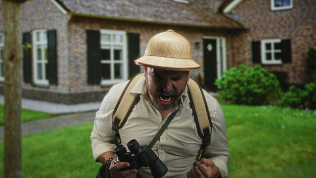 Man holding binoculars clenches fist and shouts at building entrance, backpack visible; anger exploration.
