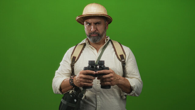Man with grey beard holding binoculars and backpack, wearing pith helmet and bracelets against a vivid green studio wall; curiosity travel exploration. - Powered by Adobe