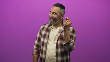 Fototapeta premium Man middle aged with grey beard pointing finger to mouth in purple studio, smiling in plaid shirt and casual pose; playful charm.