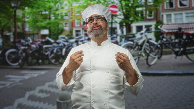 Middle aged hispanic man chef with grey beard and glasses gesturing with open hands on a street lined with bicycles and brick buildings; pride passion.