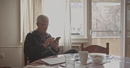 An older man in glasses sits comfortably at a table, deeply engrossed in his smart phone. He is connecting with technology, highlighting modern senior living and digital engagement at home. - Powered by Adobe