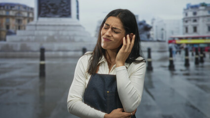 Fototapeta premium Woman holding hand to ear on wet street beside stone fountain and bollards, wearing denim apron, grimacing in pain; confusion.