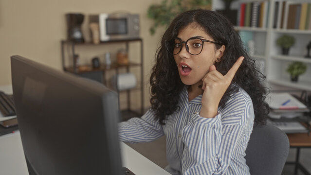 Woman resting hand on chin at office building desk, looking at computer screen while wearing glasses and a striped shirt; thoughtful pondering.