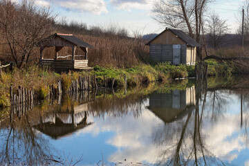 Fototapeta premium This is a rural autumn landscape featuring a calm pond or small river, with a wooden gazebo and an old hut standing on the bank, surrounded by dry grass and thickets.