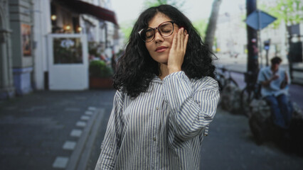 Obraz premium Woman wearing glasses touching cheek with hand on city street sidewalk near shops and bikes; urban introspection.