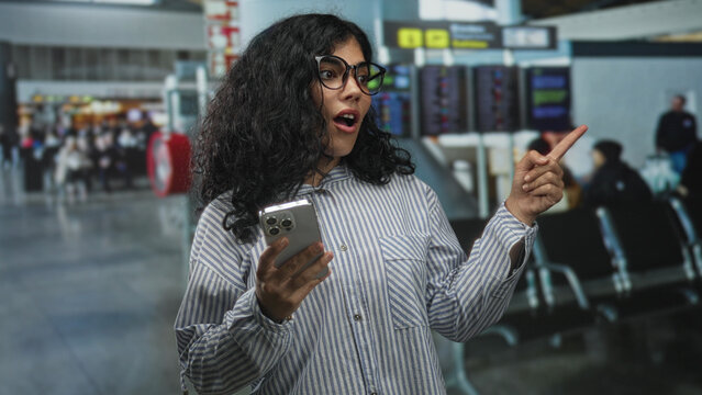 Woman holding smartphone points finger at departure board in airport terminal; anticipation travel planning.