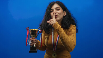 Young arab brunette woman holding trophy and wearing a number one medal, finger to lips for silence in blue studio; victory hush.