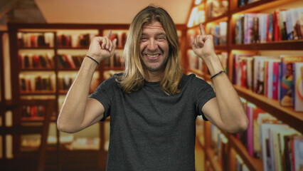 Man with long hair wearing dark shirt finger pointing to books on shelf in library while smiling broadly; excitement.